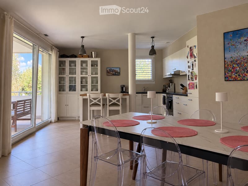 Open-plan kitchen and dining area with a large central dining table, modern white cabinets, and a view to a balcony.
