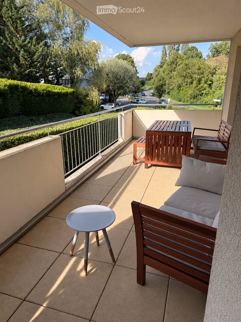 Balcony with a wooden sofa, a small round table, and a wooden chair, overlooking a tree-lined street.
