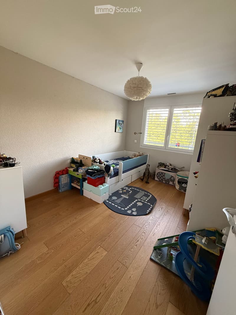Child's bedroom with wooden flooring, a white bed with drawers, a colorful table, a blue chair, toys, and a round rug with a road design.