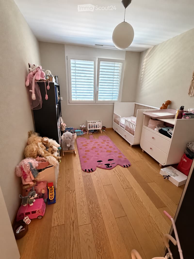 Brightly lit children's room with wooden flooring, crib, changing table, wardrobe, and various toys including a plush elephant, toy car, and a changing mat.