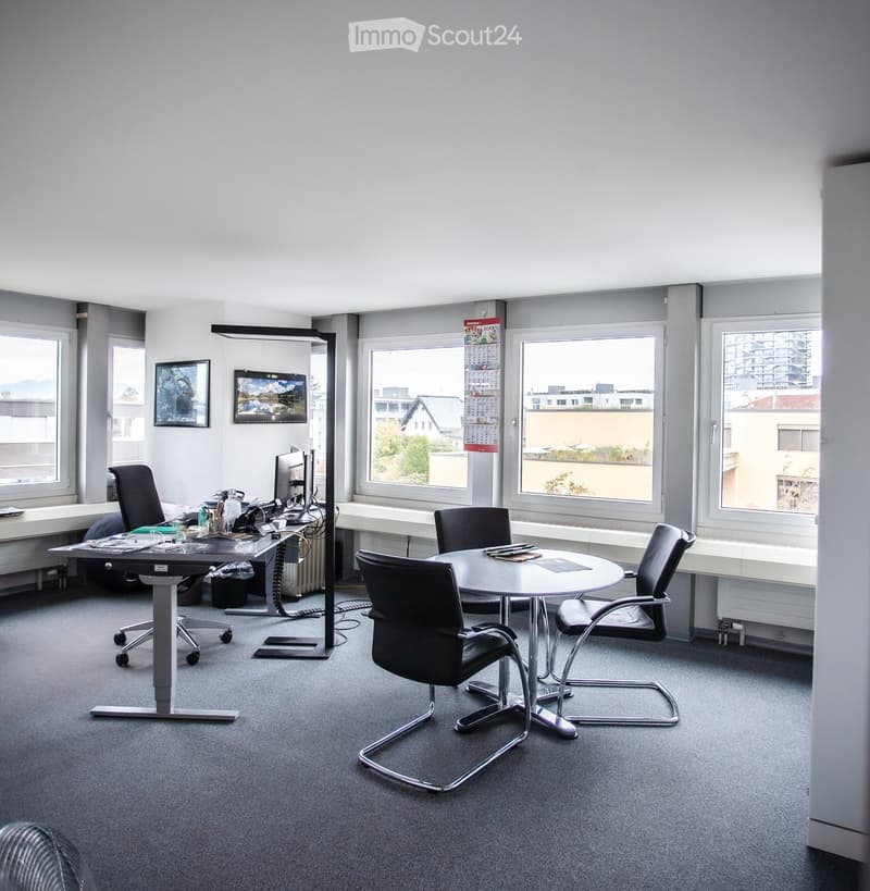 Office space with a round meeting table, four chairs, a standing desk with a computer, and a floor lamp. Large windows provide natural light.