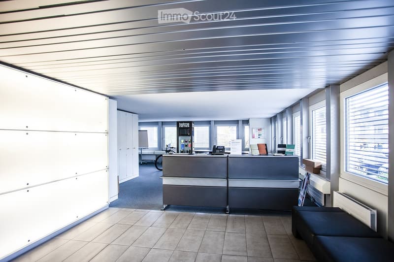 Modern office space with a reception desk, sofa, laptops, and a cabinet. The room features large windows and a tiled floor.