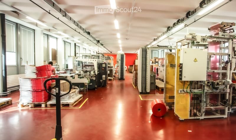 Industrial factory space with red flooring, large windows, and various machinery and equipment, including stacked red and white materials on pallets.