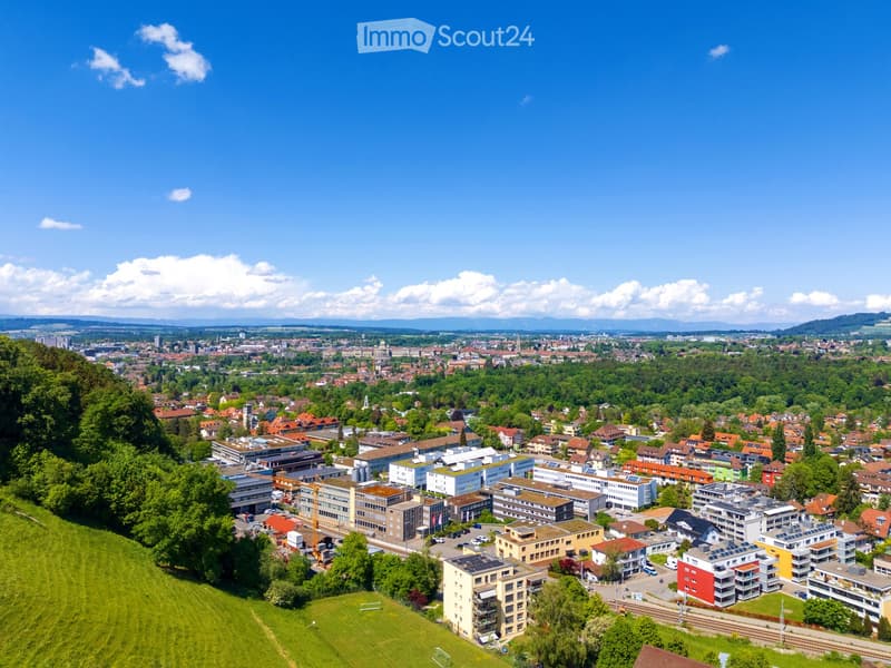 Aerial view of a town with a mix of residential and commercial buildings, surrounded by green spaces and trees, under a clear blue sky.