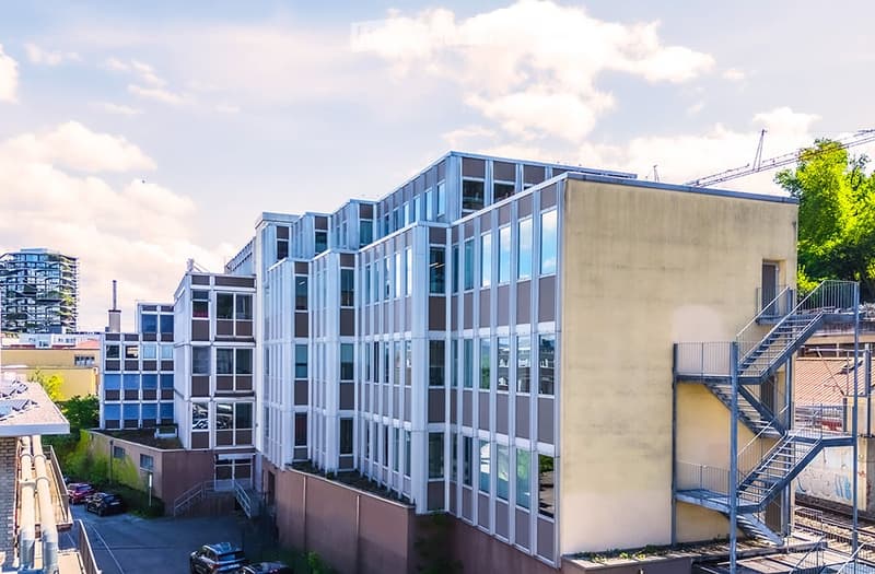 Modern multi-story building with large glass windows and a mix of beige and brown facades, surrounded by other structures and parked cars.