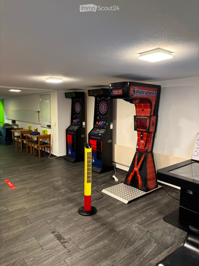 A recreational room with a dartboard, arcade machine, and a Spider-Man themed game. There is a small table and chairs in the background.