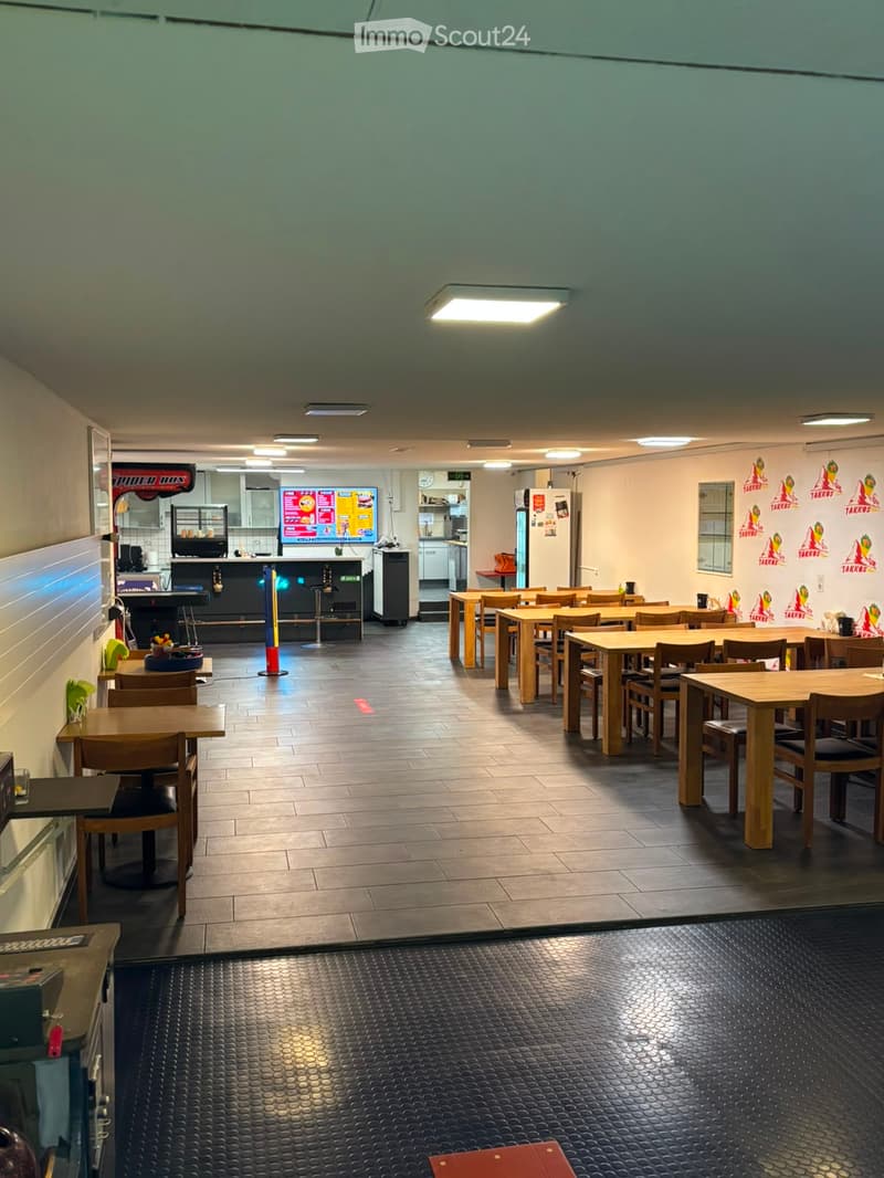Restaurant interior with multiple tables and chairs arranged in rows, a counter area with stools, and a visible kitchen area with appliances and a refrigerator.
