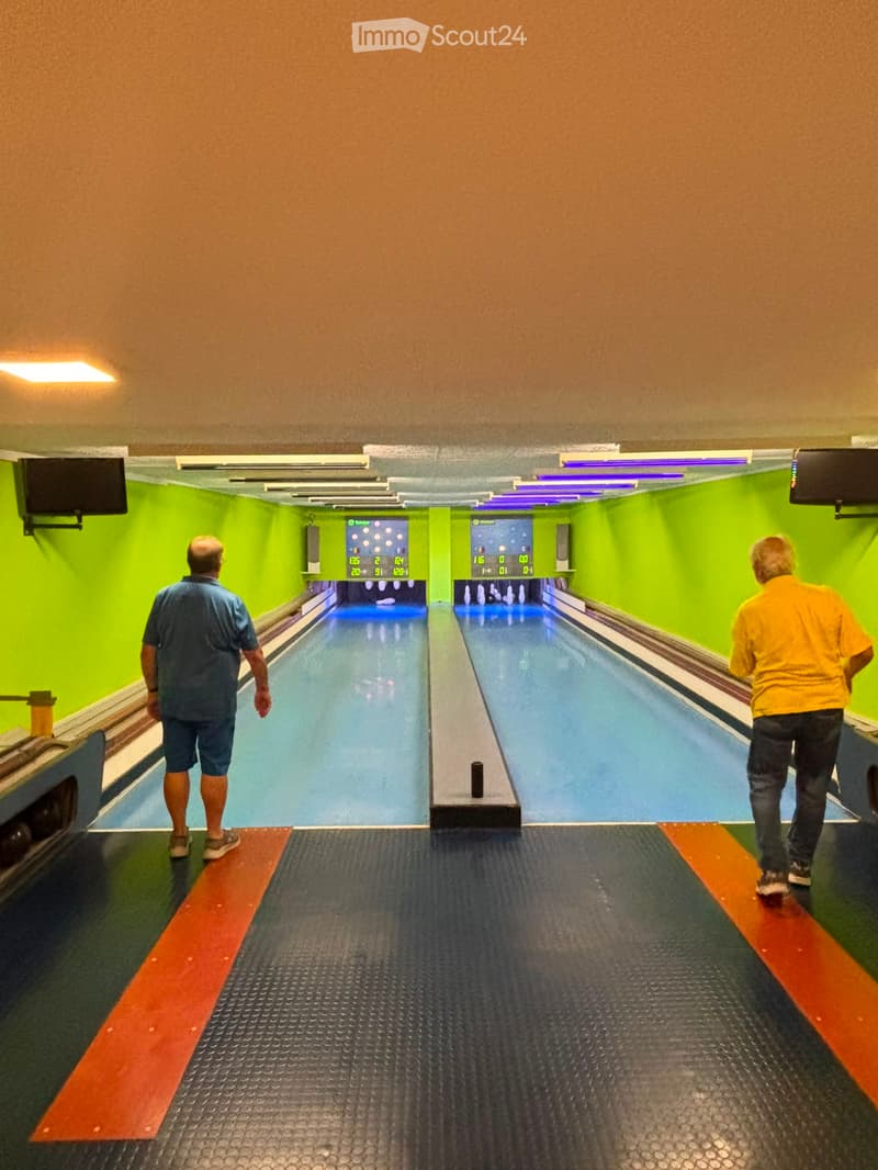 Bowling lanes with two lanes visible, each with pins at the end and a digital scoreboard above. Two individuals are walking towards the lanes.