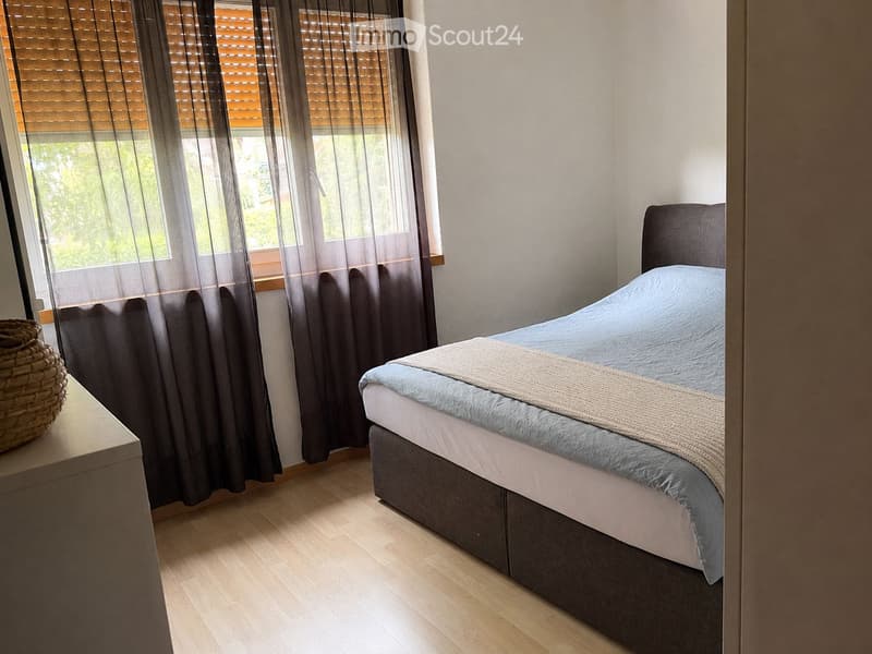 A modern bedroom with a neatly made bed, a nightstand with a wicker basket, and large windows with dark curtains and blinds.