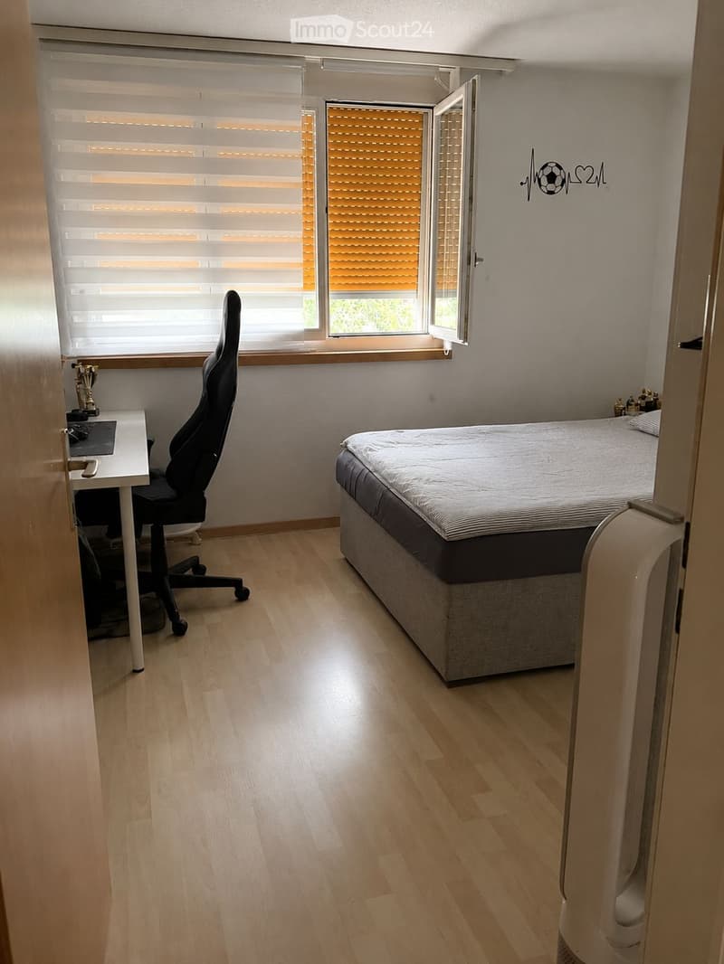 Modern bedroom with wooden flooring, a double bed, a desk with a laptop, and a black office chair. The room features large windows with blinds and a small air conditioning unit on the right side.