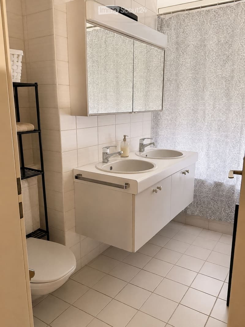 Modern bathroom with white tiles, double sink vanity, mirrored cabinet, and a toilet. The shower area features a decorative curtain.