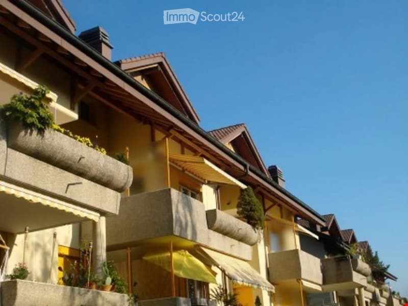 Multi-story residential building with balconies, wooden awnings, and potted plants. The structure features a combination of yellow and beige facades with a traditional roof design.
