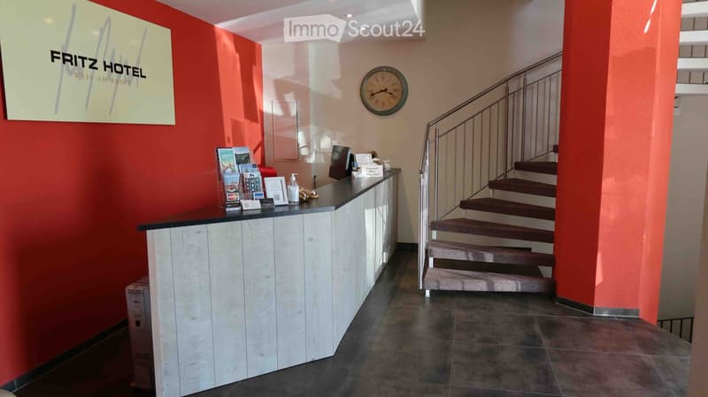 Hotel lobby area with reception desk, staircase, clock, and informational materials.