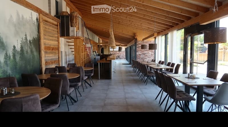 Restaurant interior with wooden tables, chairs, and pendant lights. Features a staircase leading to an upper level and a mural of trees on one wall.