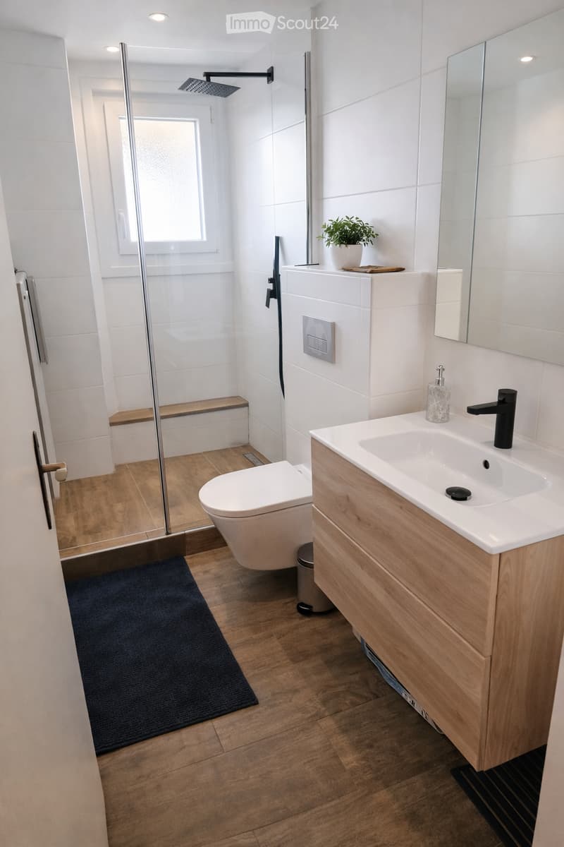 Modern bathroom with wooden flooring, white tiles, a toilet, a sink with a wooden cabinet, a shower area with a glass door, a mirror, and a small plant on the cabinet.