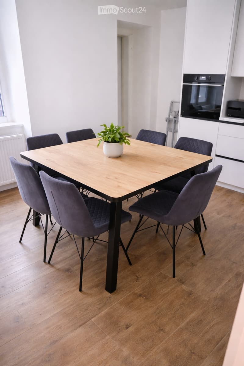 Dining area with a wooden table, six dark blue chairs, and a potted plant on the table. The floor is wooden, and the room has white walls and a radiator visible on the left side.