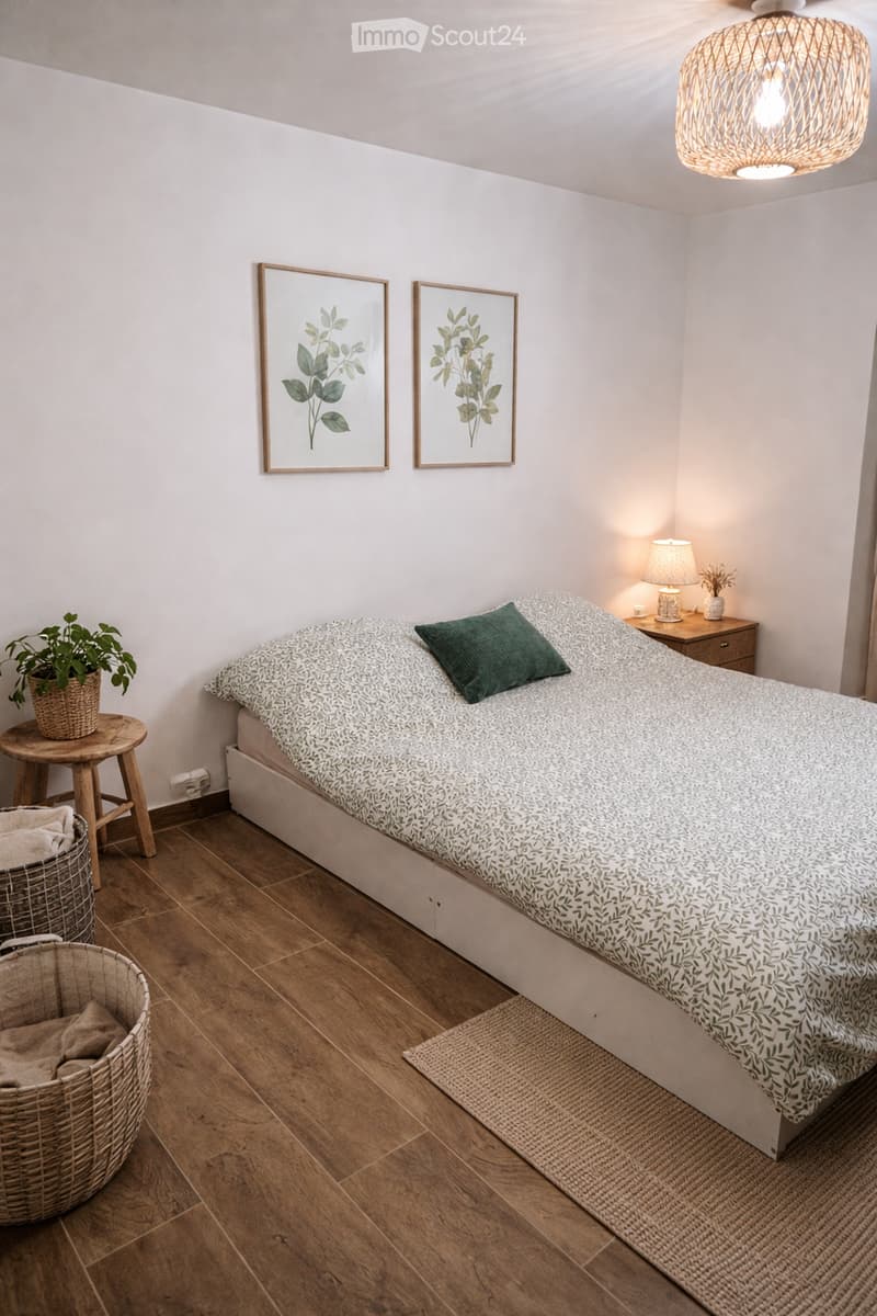 Cozy bedroom with a white bed, green pillow, nightstand with lamp, small wooden stool with plant, and two framed botanical prints on the wall.