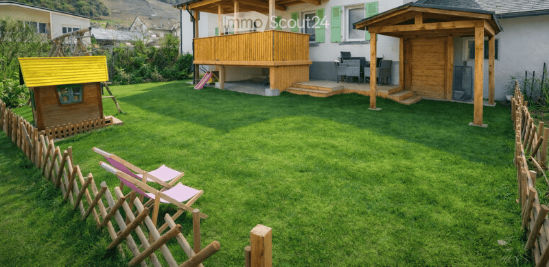Well-maintained garden with green grass, wooden fence, pink folding chairs, small playhouse, wooden deck, and outdoor seating area.