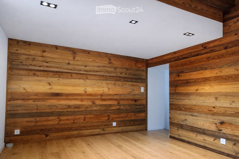 Empty room with wooden plank walls and floor, recessed ceiling lights, and a small open doorway leading to another space.