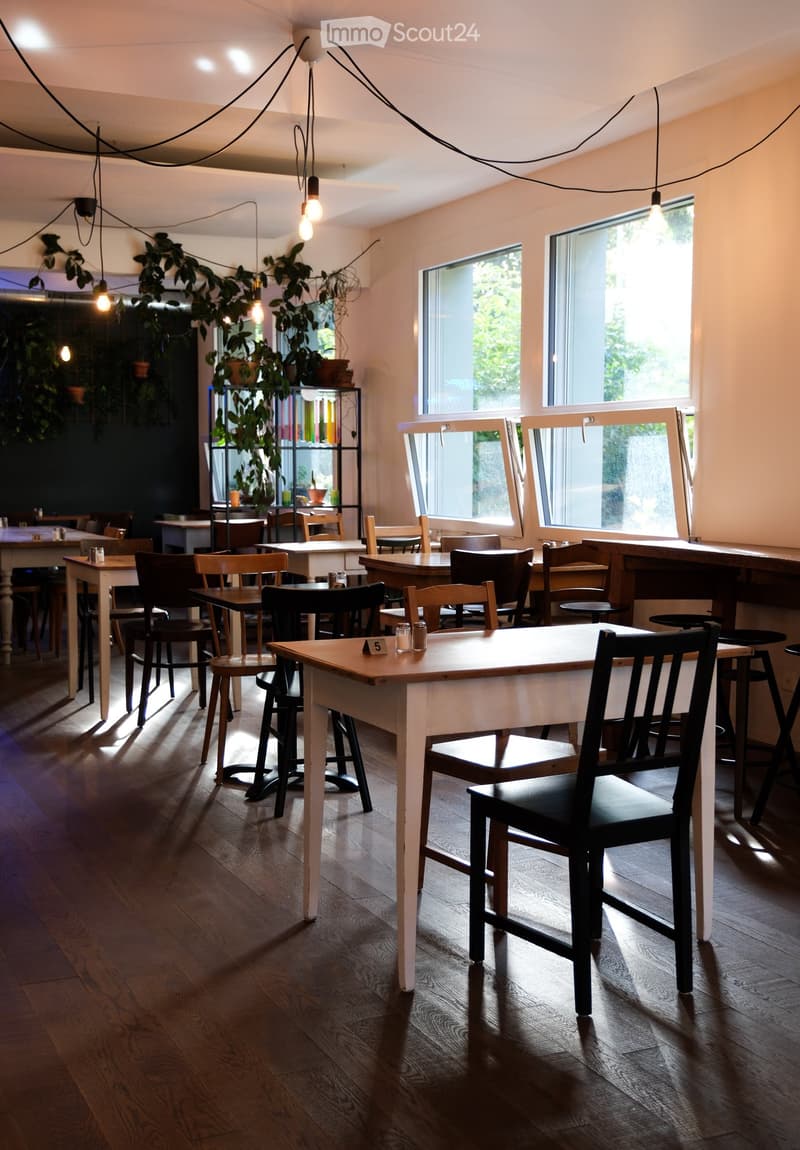 Brightly lit dining area with wooden tables, chairs, and stools arranged around the space. Large windows allow natural light to fill the room, complemented by hanging pendant lights. Potted plants and shelves add a touch of greenery and decor.
