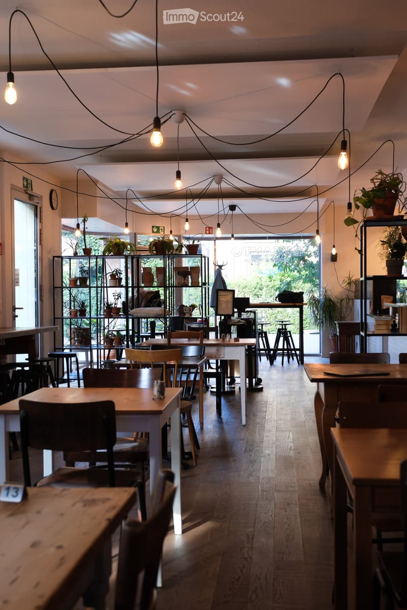 A modern, well-lit restaurant with wooden tables and chairs, hanging pendant lights, and shelves with potted plants and decorative items.