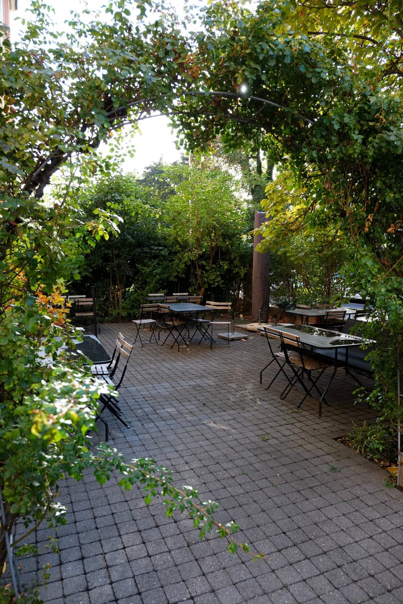 An outdoor seating area with multiple tables and chairs arranged on a paved surface, surrounded by lush greenery and trees.