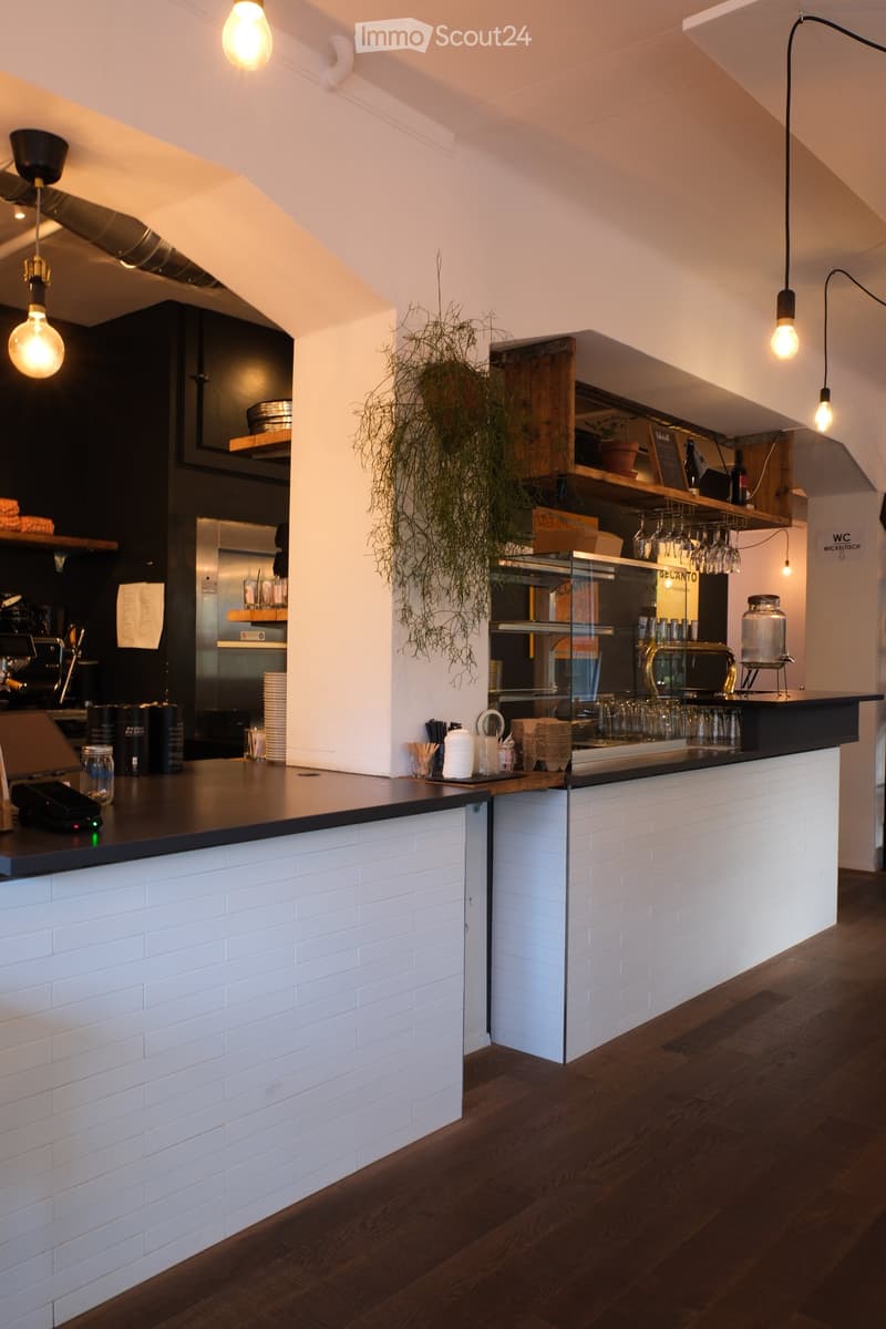 Modern bar area with white brick walls, dark wooden floor, and a long counter. Features pendant lights, wine glasses, a potted plant, and various bar accessories.