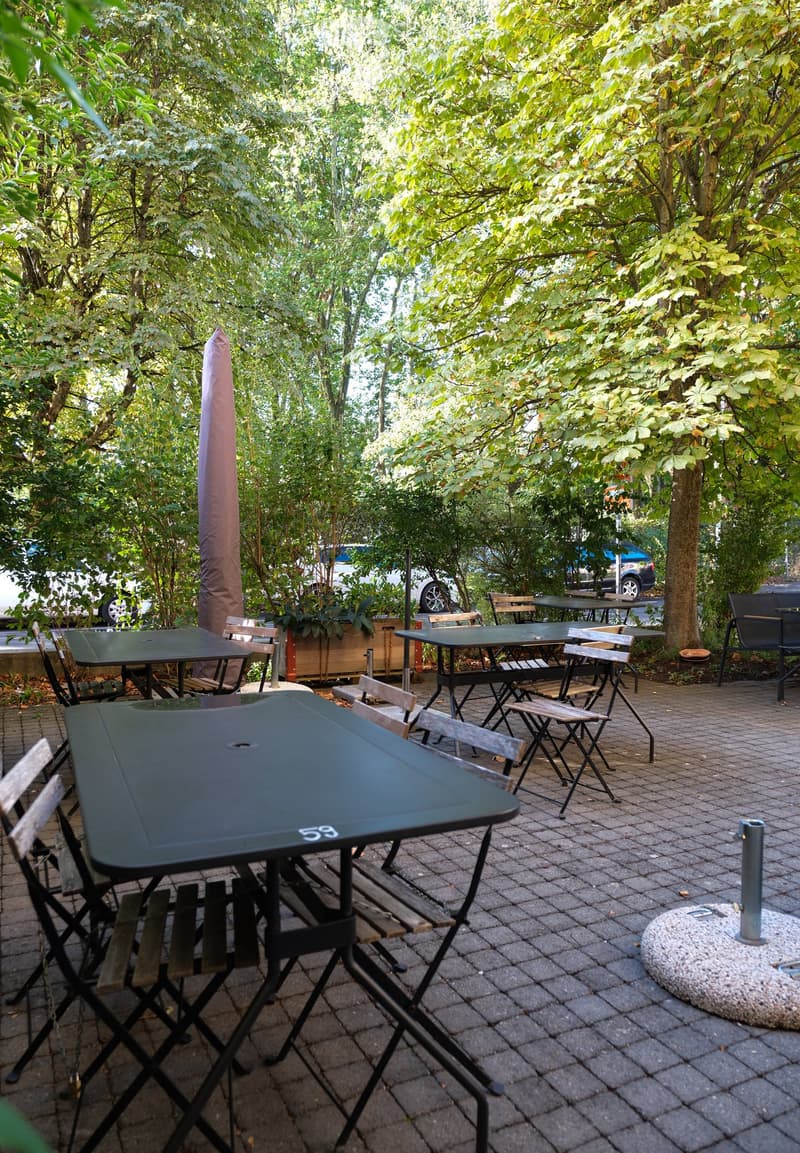 An outdoor seating area with several tables and chairs arranged on a paved surface, surrounded by lush greenery and trees.