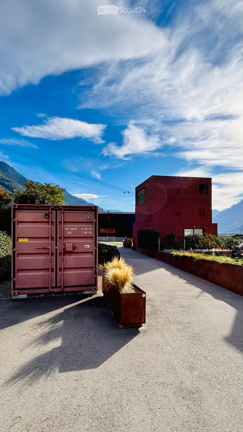 Outdoor area with a paved pathway, a large red shipping container, a planter with tall grass, and a modern red building in the background, surrounded by greenery and mountains.