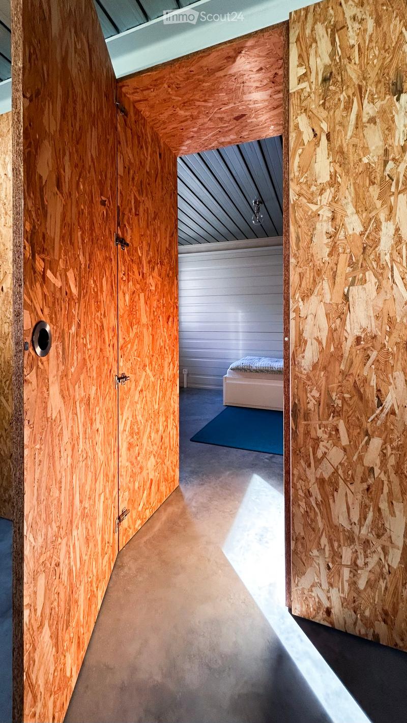 A modern hallway with wooden panel walls, a concrete floor, and a visible bedroom area with a bed and rug through an open doorway.