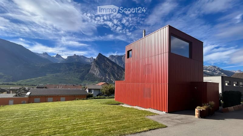 Modern red metal house with large windows, surrounded by a green lawn and mountains in the background.