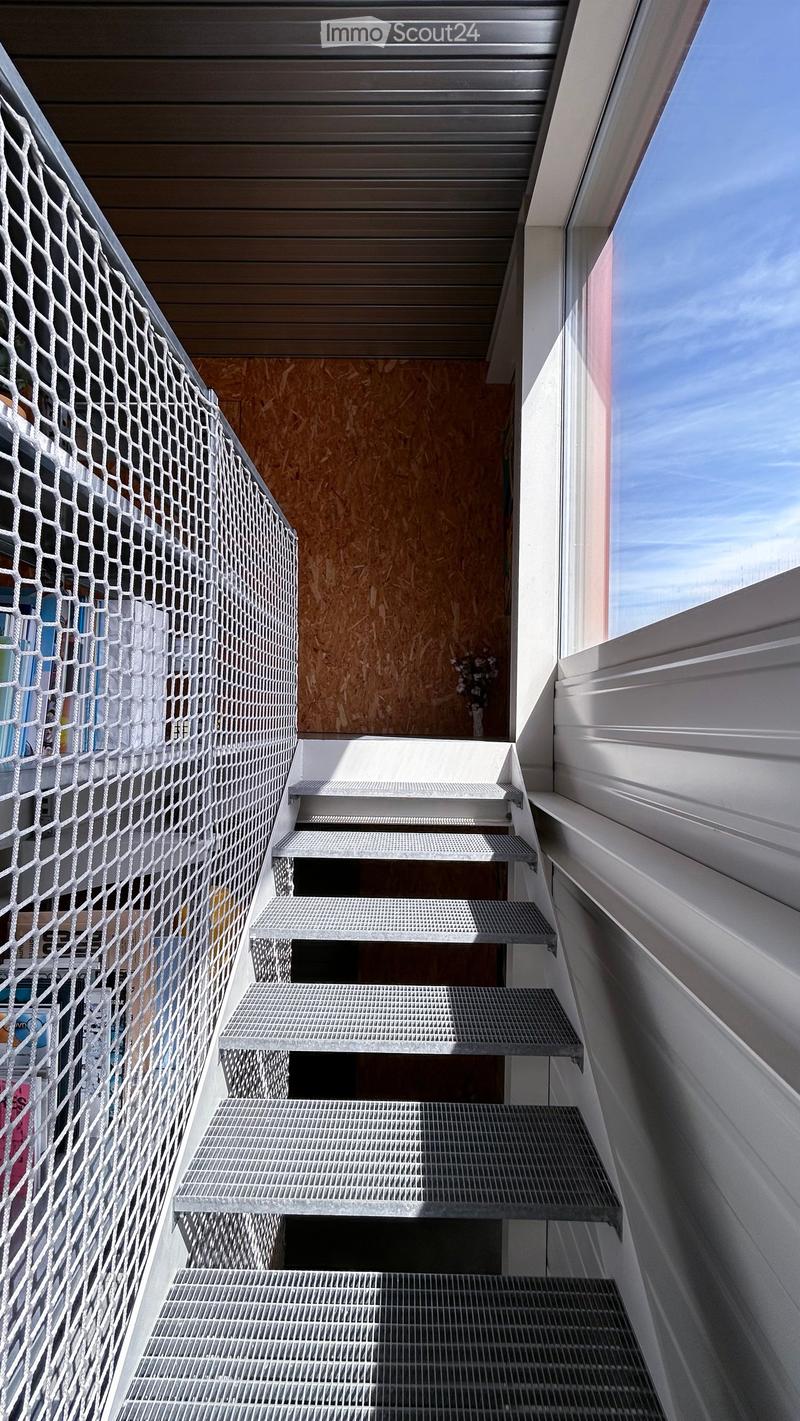 Metal staircase with a wire mesh railing, leading up to a wooden door with a small window and a vase of flowers on a shelf.