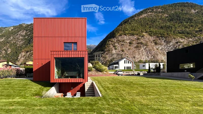 Modern red house with large windows and a balcony, set in a grassy yard with a mountain view in the background.