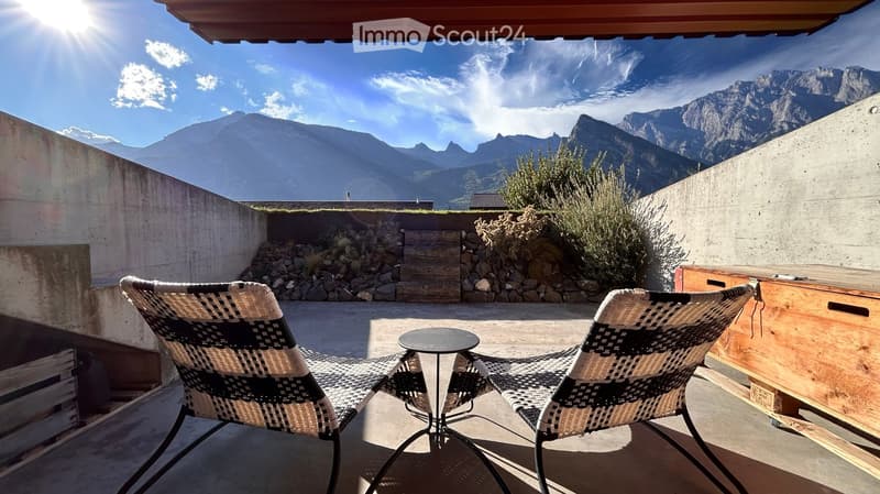 A modern outdoor terrace with two wicker chairs and a small round table, overlooking a scenic mountain view.