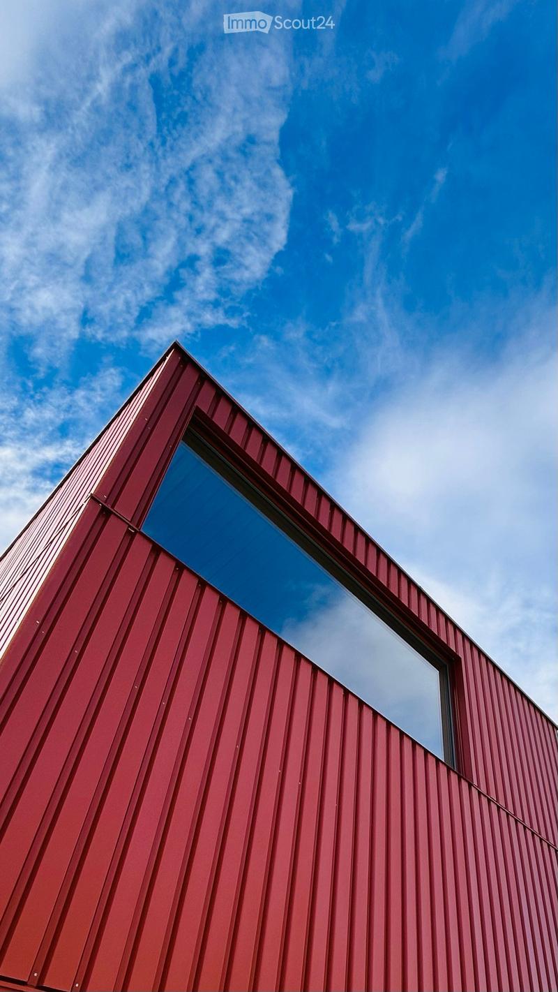 A modern red metal building with a large, reflective window on its side, set against a clear blue sky with scattered clouds.