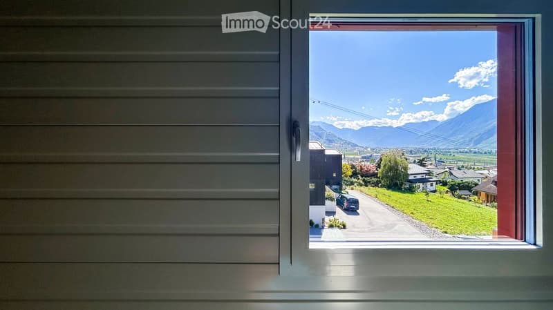 View from a window showing a scenic landscape with mountains, a road, and houses in the distance.