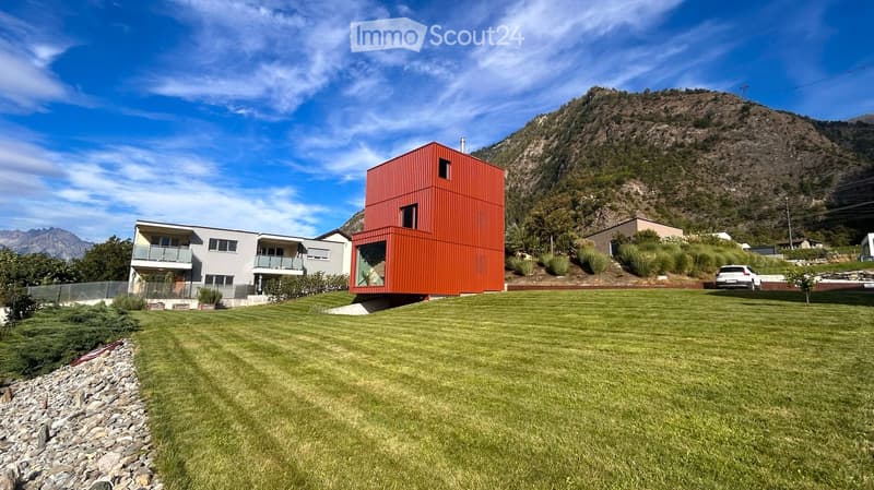 Modern two-story house with a red container-style extension, surrounded by a well-maintained lawn and set against a mountainous backdrop.