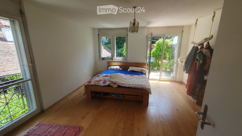 Bright bedroom with wooden flooring, large windows, and a bed with colorful bedding. A coat rack with jackets is visible near the door.