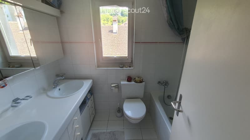 A clean, modern bathroom with a white sink, toilet, bathtub, and a large mirror above the sink. The room features tiled walls and a window with a view of the roof.