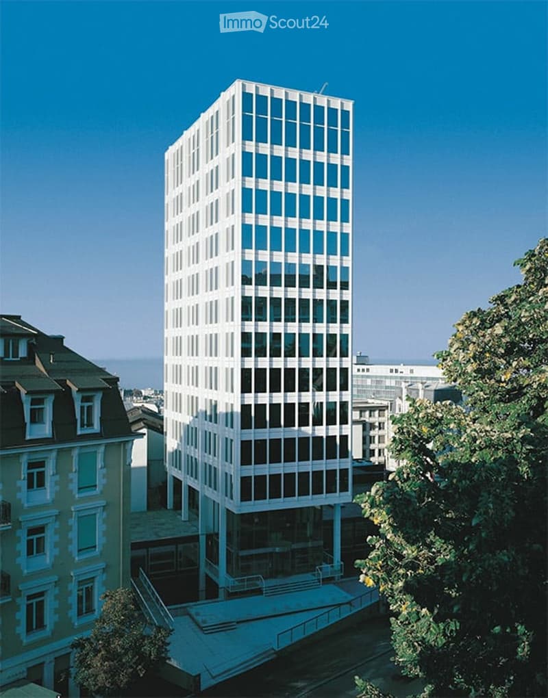 Modern high-rise building with a grid of windows, surrounded by smaller structures and greenery.
