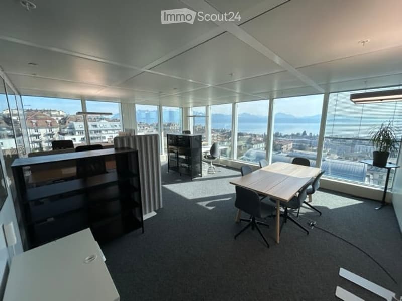 Modern office space with large windows offering a scenic view, featuring a wooden table, chairs, a cabinet, and a potted plant.