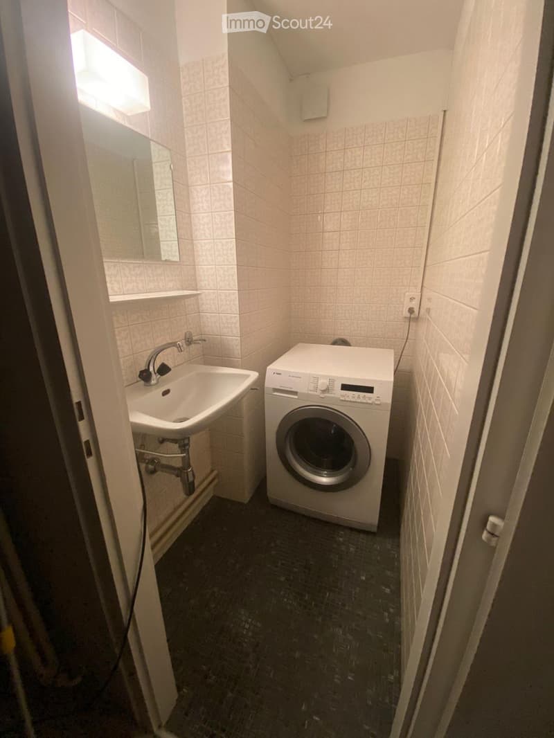 Small bathroom with a sink, faucet, washing machine, and mirror. The walls are tiled, and the floor is dark.