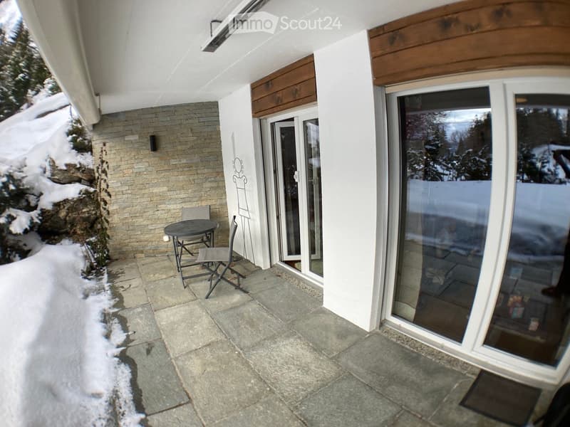 Balcony with a small table and two chairs, snow-covered ground, large glass doors leading inside, brick wall, and wooden trim.