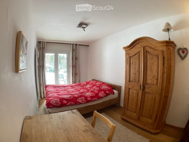 Cozy bedroom with a wooden bed, red patterned bedspread, large wooden wardrobe, small wooden table with a chair, and a lamp on the wall.