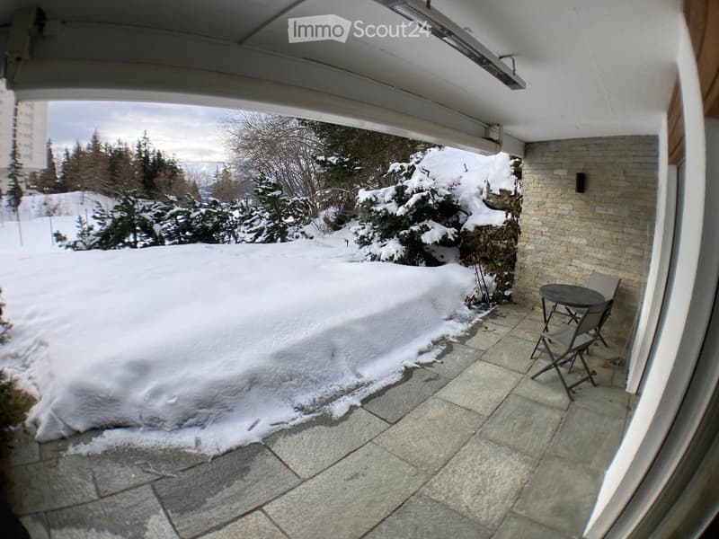 Balcony with stone tiles, snow-covered ground, small table and chairs, surrounded by snow-covered trees and a brick wall.