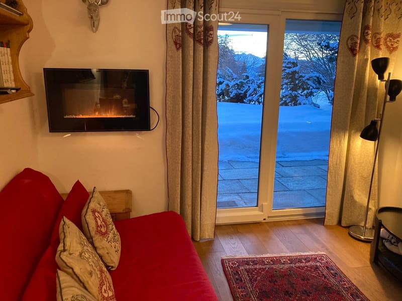 A cozy living room with a red sofa, decorative pillows, a wall-mounted fireplace, a floor lamp, a small table, and a shelf. Large windows offer a view of a snowy outdoor scene.