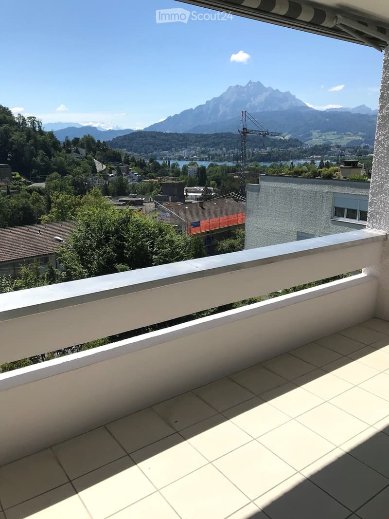 Balcony with tiled floor and white railing, overlooking a scenic landscape with mountains, trees, and a body of water.