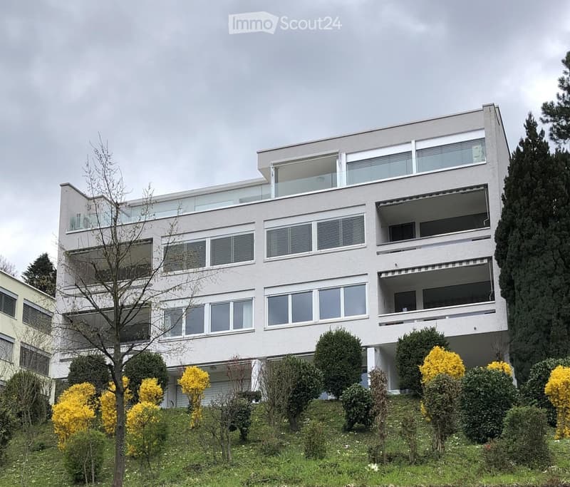 Modern multi-story building with large windows and balconies, surrounded by well-maintained greenery and shrubs.