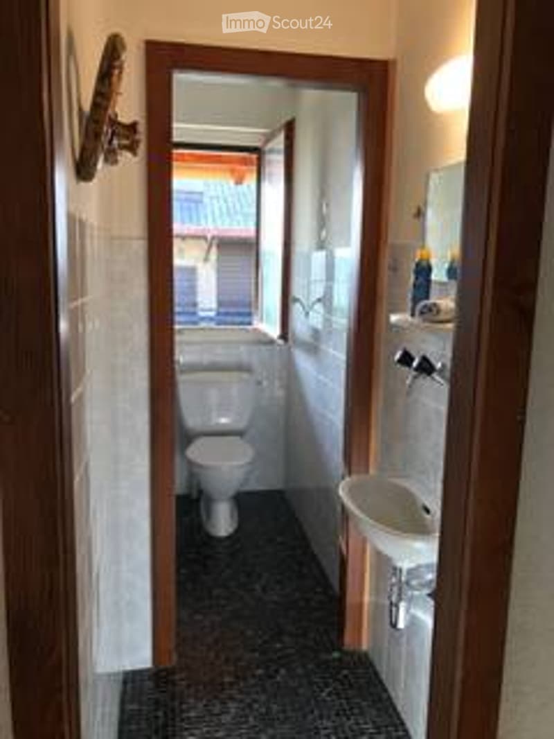 Small bathroom with white tiled walls, black tiled floor, toilet, sink, and mirror. The sink has a modern faucet and a small shelf above it holding a bottle of cleaning supplies.