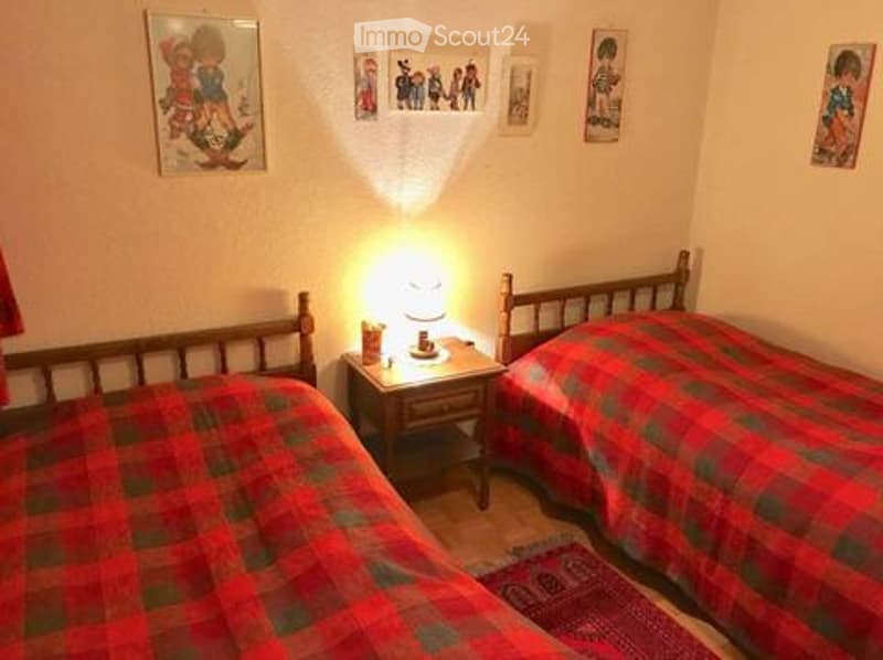 Cozy bedroom with two single beds, red and green checkered bedding, a wooden nightstand with a lamp, and several picture frames on the walls.
